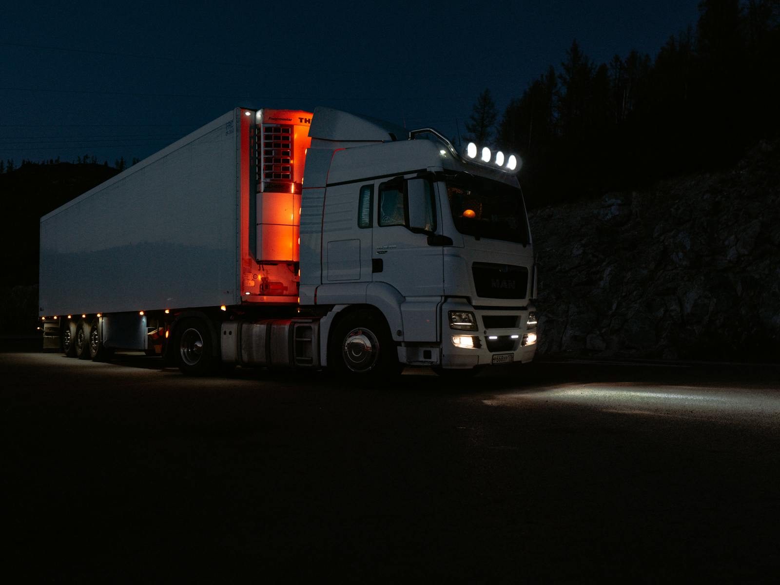 A white truck lit by headlights and interior lights on a dark road in mountainous terrain.